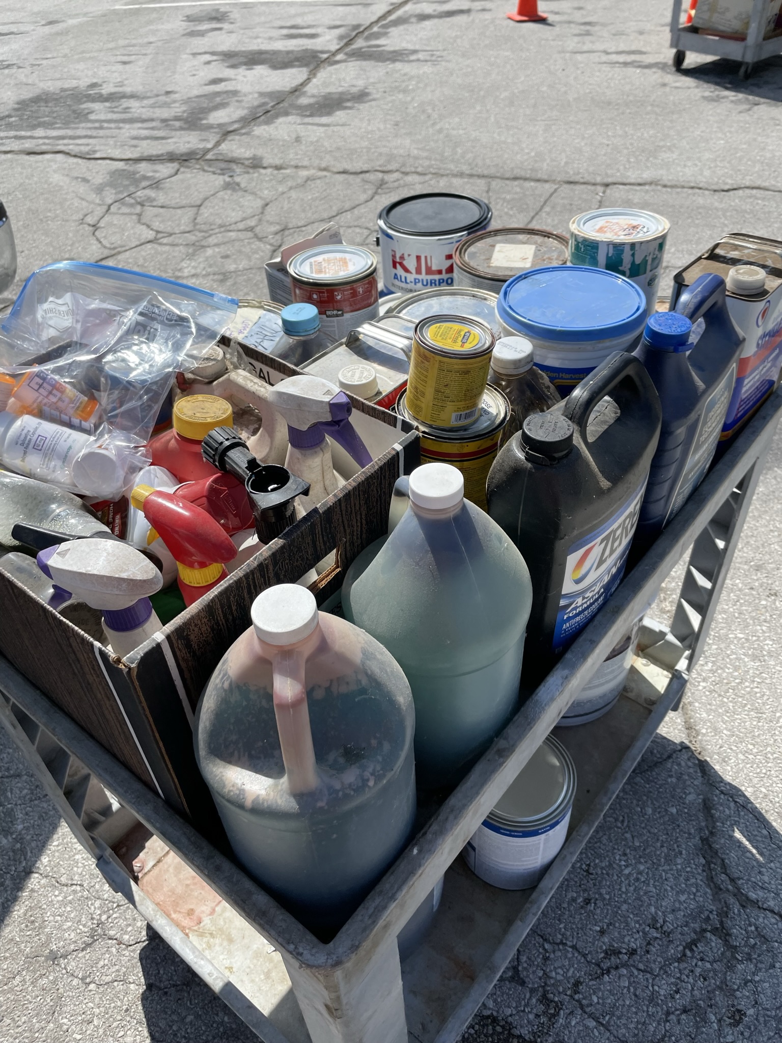 A cart filled with old paint and other household hazardous waste items sits in the sunshine in a parking lot waiting to be disposed of safely. 