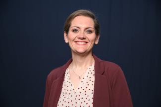 A portrait image of Samantha Morris in a maroon jacket with a peach and maroon shirt under it in front of a dark blue background.