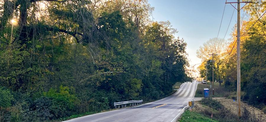 Straight paved road surrounded by trees and power lines on the right, with newly installed guardrails and a fresh pave job over a culvert that was damaged by flooding rains.