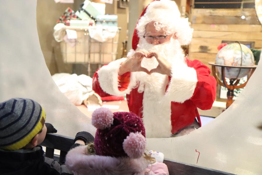 Santa in window making heart with hands. Children outside looking in window at Santa.