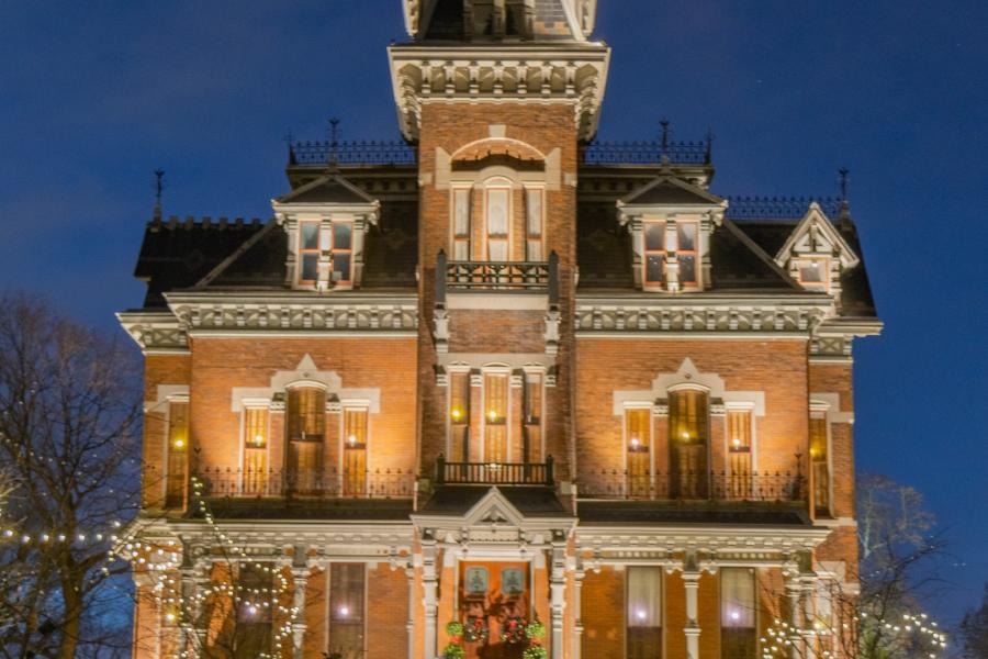 Image of the outside of the Vaile Mansion with holiday lights on the mansion and shrubs. 