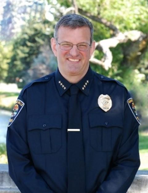 Chief Bryce Johnson, a candidate for the Independence Chief of Police, stands in uniform in a nature setting with trees, a river, and mountains behind him.