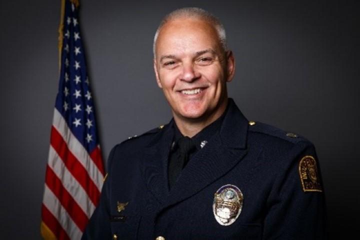 Deputy Chief Ken Jarnagin smiles in an official portrait wearing his police uniform with an American flag behind him and a dark grey background.