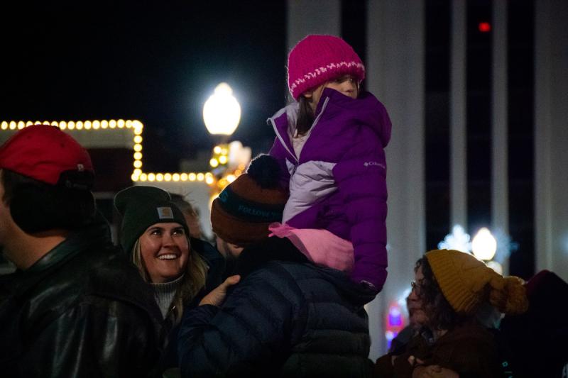 Image of a girl on father's shoulders. Woman looking up at girl and smiling. 