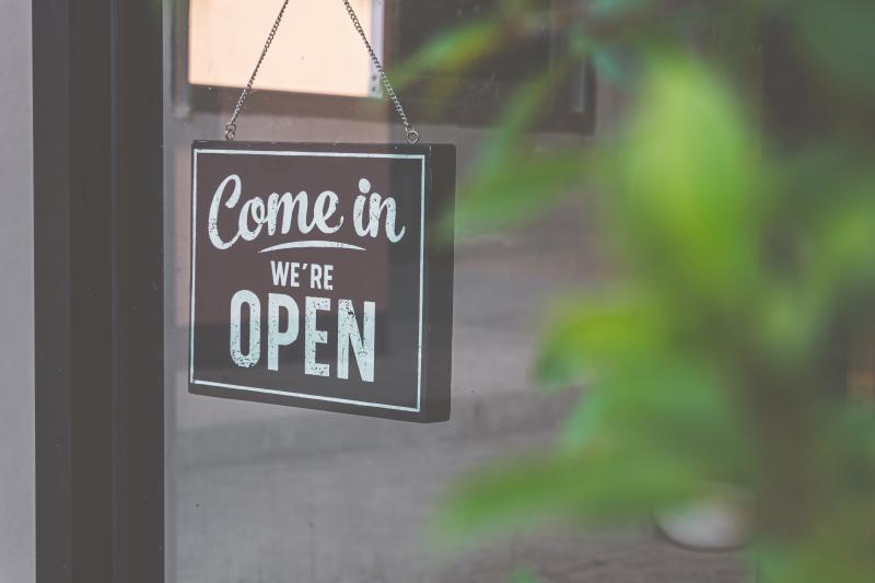 Come in we're open sign hangs in the window of a storefront with greenery behind it. 