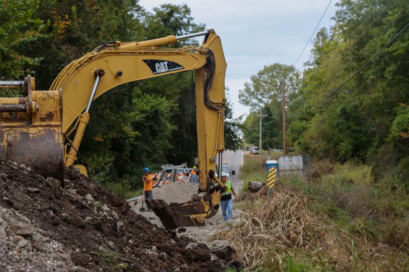 A backhoe in the foreground, digging up the ground while construction workers look on